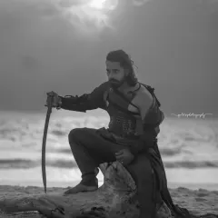 A black and white shot from a warrior-themed editorial on the beach. The lack of color enhances the dramatic sky and the rugged texture of the scene, focusing on the subject's powerful silhouette.