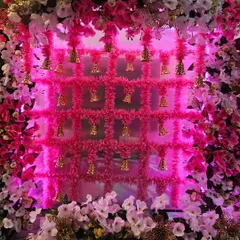 A vibrant and festive backdrop featuring a grid of pink tassels and golden bells, framed by pink and white flowers. This piece is perfect for Navratri or a baby shower and is available to book now.