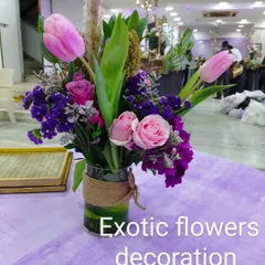 A modern table centerpiece for an event, featuring pink tulips, roses, and purple statice in a glass vase wrapped with jute rope. This style is perfect for corporate events or contemporary celebrations.
