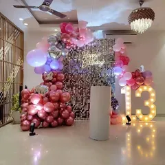 A trendy 13th birthday party with a rose gold shimmer wall, a 'Happy Birthday' neon sign, and a '13' marquee light, framed by a matching balloon garland.
