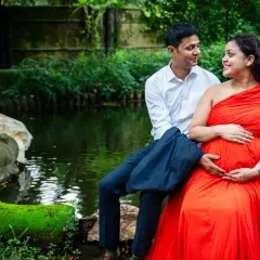 A quiet, candid moment shared between an expecting couple by the water. The vibrant red of her dress stands out beautifully against the serene, green landscape of the park.
