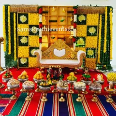 A classic baby shower setup with a checkerboard backdrop of yellow marigolds and woven green leaves. The traditional sofa and a full display of offerings on a striped mat complete this authentic South Indian look.