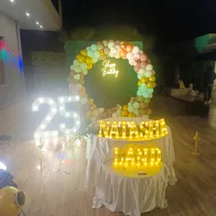 A stylish outdoor 25th birthday party setup. The decoration features large, illuminated marquee numbers for the age, a custom name sign in marquee letters, and a circular balloon arch against a green foliage wall.