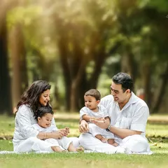 A family with twin boys enjoying a picnic-style setup in a park. The all-white outfits create a classic, clean look against the green grass.