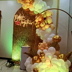 A glamorous engagement photo corner. The backdrop features a gold sequin panel, a "Better Together" neon sign, and an organic arrangement of gold, white, and yellow balloons.