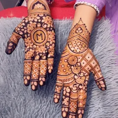 A festive Diwali mehendi with the initial 'M' in the center. One hand has a dense floral pattern, while the other has a more geometric design.