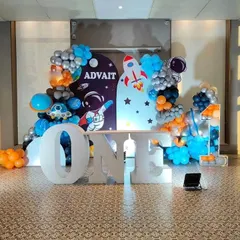 An out-of-this-world space theme for a first birthday, with large "ONE" marquee letters in the foreground. The backdrop features astronauts and rockets, framed by a blue, orange, and silver balloon arrangement.