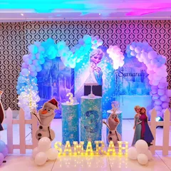 An enchanting Frozen birthday theme with a shimmering ice castle backdrop. The setup features life-size cutouts of Elsa and Anna, along with a blue and white balloon arch.
