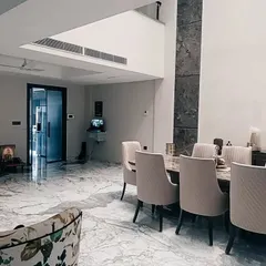 In this Gurgaon residence, the dining area flows into the living space, unified by expansive marble flooring. The design emphasizes verticality with a tall, stone-clad feature wall, creating a sense of grandeur in the home.