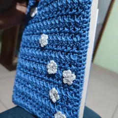 A close-up of my blue crochet book cover, adorned with small white flowers. It's designed to protect your pages while adding a touch of handmade charm.