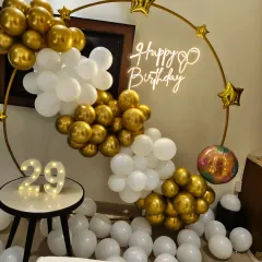 A stylish 29th birthday party setup. The gold and white balloon garland on a ring frame, paired with a neon sign and marquee numbers, creates a perfect photo opportunity.