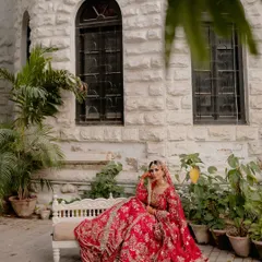 A moment of quiet contemplation for the bride. Seated gracefully, her vibrant red lehenga spread around her, she looks like royalty against the rustic charm of the stone building.