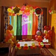 A unique and colorful fruit-themed birthday party. This vibrant setup includes a multi-colored fringe backdrop, fruit-shaped foil balloons, and a bright neon 'Happy Birthday' sign.