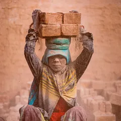 A woman, her face and clothes covered in dust, sits with a load of bricks on her head, her direct gaze telling a story of resilience.