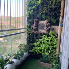 The same balcony garden viewed during the day, showcasing a mix of plants like Syngonium and Spider Plants. The design combines a vertical wall with potted plants and artificial grass to create a complete, low-maintenance oasis.