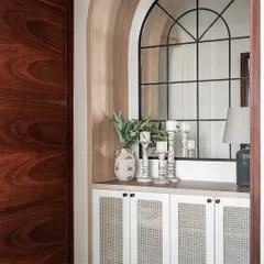 An elegant corridor with a custom-built storage cabinet featuring woven rattan doors. The arched mirror and dark wood door create a beautiful contrast and a sense of classic charm.