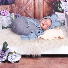 A cozy setup featuring a baby in a cute mouse hat, sleeping on a fluffy bed with a matching teddy bear. The lavender floral background complements the grey wrap.
