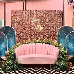 A chic and modern photo booth for a party. It features a rose gold sequin wall, a "You & Me" neon sign, and tropical leaf panels, all centered around a stylish pink sofa.