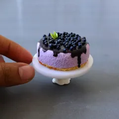 A close-up of a miniature blueberry cheesecake, showing the glossy topping and crumbly base, all made from air-dry clay.