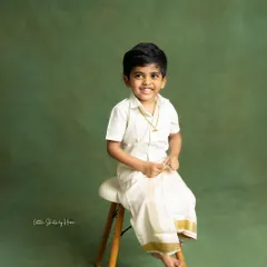 A handsome little boy dressed in a traditional veshti and shirt. This simple, classic portrait against a solid green backdrop highlights his bright smile and cultural attire.