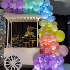 The white wooden candy cart, decorated with a cascade of rainbow balloons, serves as a beautiful and functional piece of decor. It can be used to display sweets, gifts, or a cake.