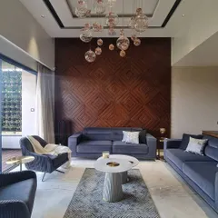 In this living room, we created a feature wall with an intricate geometric pattern in dark wood, adding warmth and texture to the space. The design is complemented by modern blue sofas, marble coffee tables, and a cluster of glass globe pendant lights.