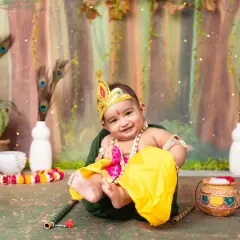 A joyful baby boy dressed as Krishna, with a big smile, sitting in a green bowl amidst a festive setup.