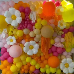 A vibrant and textured balloon wall made from a mix of yellow, pink, and orange balloons. The addition of daisy-shaped balloons and tassels adds a fun, artistic touch to the installation.