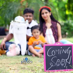 A family maternity shoot in a park, announcing their second child with a "Coming Soon" sign and including the older sibling in the portrait.