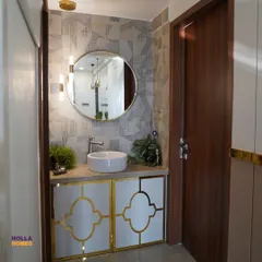 An elegant basin area design that makes a statement of luxury. It features gold-accented cabinets, a round mirror, and a geometric-patterned wallpaper.