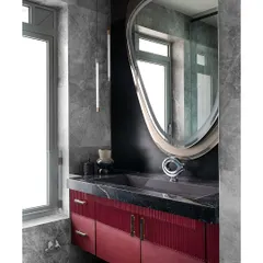 In this master bathroom, a rich red vanity with fluted detailing adds a pop of color against the grey marble walls. The organically shaped mirror and sleek pendant light enhance the modern, luxurious feel of the space.