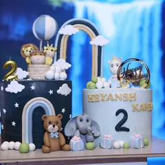 A set of two coordinated cakes for a second birthday party. One cake features a starry night theme with a teddy bear and elephant, while the other has a hot air balloon with a lion and giraffe.