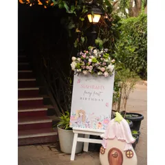 The entrance to the fairy party, featuring a custom welcome sign on an easel, topped with a beautiful floral arrangement and flanked by a whimsical mushroom house prop.