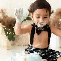 A playful moment from a boy's cake smash session, showing off his frosting-covered hands against a stylish boho backdrop.