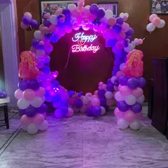 A beautiful ring-shaped balloon arch in shades of purple, pink, and white. The neon "Happy Birthday" sign in the center adds a modern and vibrant touch, perfect for a stylish home party.