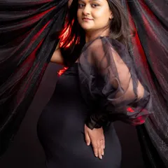 A classic maternity pose against a dark, textured backdrop. The expecting mother, in a fitted black dress with sheer sleeves, gently holds the fabric, looking confidently at the camera.