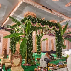 A traditional South Indian mandap setup inside a hall, featuring pillars wrapped in greenery and banana plants.