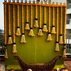 A beautiful and traditional backdrop design suitable for a Mehndi or Haldi function. The base is a simple green cloth, which makes the hanging garlands of marigolds and white jasmine flowers pop, creating a fresh and festive look.