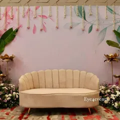 This beautiful backdrop was designed for an engagement party, featuring a soft, floral-printed background with hanging floral strings. The setup is framed by tropical leaves and flower arrangements, with a plush beige sofa for photos.