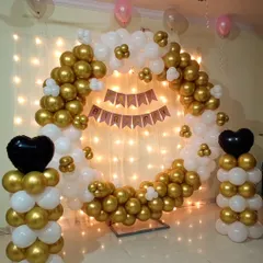 This birthday decoration features a circular balloon ring in gold and white, set against a wall of fairy lights. The design includes balloon pillars topped with black heart foil balloons for a touch of contrast.