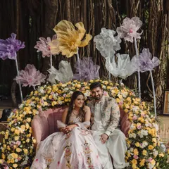 A dreamy Mehendi seating area. The couple is framed by a crescent of yellow and pink flowers, with whimsical, oversized paper flowers adding a touch of magic to the natural backdrop.