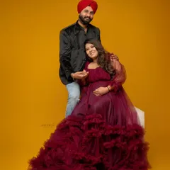 A full-length studio portrait of an expecting couple. The wife is seated in a stunning ruffled magenta gown, showcasing a popular style for modern maternity photoshoots.