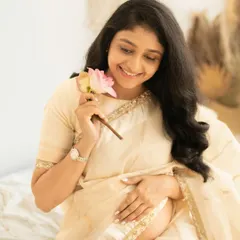 A sweet and delicate portrait of a mother-to-be in a cream saree. She is smiling as she holds a single pink lotus flower, captured in a soft and dreamy boho setting.