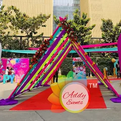 A vibrant and colorful entrance designed for a corporate Holi party. The setup features a red carpet walkway under a canopy of bright ribbons and flanked by large, playful, multi-colored arches, setting a fun and festive tone from the start.