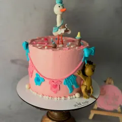 A sweet baby shower cake with a gender-neutral theme. It features a fondant stork topper and a clothesline with tiny pink and blue baby clothes.
