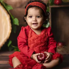A happy baby girl in a bright red dress smiles for the camera, holding an apple in a rustic, themed studio setup. Her cheerful expression is absolutely infectious.