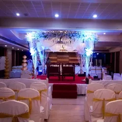 An elegant indoor mandap setup in a banquet hall. The design uses white drapes, a floral canopy, and coordinated chair covers to create a cohesive and beautiful ceremony space.