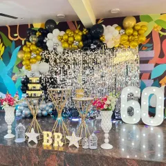 A glamorous 60th birthday party setup. The decor features a silver shimmer wall, a black and gold balloon arch, and large light-up marquee numbers.