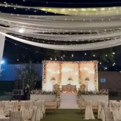 A wide view of an outdoor reception setup at night. The elegant white stage is complemented by a ceiling of fairy lights and drapes, creating a magical ambiance.