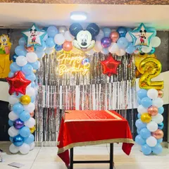 A Mickey Mouse themed birthday party setup. It includes a balloon arch with a Mickey Mouse head, a 'Happy Birthday' neon sign, and the number '2' in a large foil balloon.